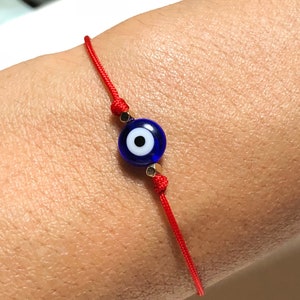 Puede incluir: Pulsera de hilo rojo con una cuenta de ojo turco azul y blanco. La pulsera tiene cuentas doradas a ambos lados del ojo. La pulsera está atada con un nudo corredizo para un ajuste ajustable.