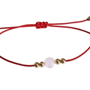 May include: Red string bracelet with a central pink gemstone and gold-colored beads. The adjustable bracelet features a sliding knot closure and small gold beads at the ends of the string.