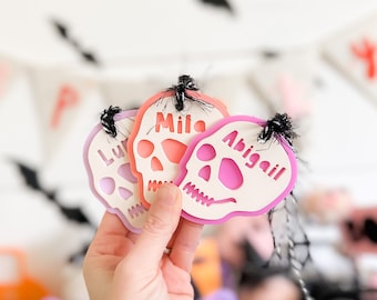 May include: Three pink and purple Halloween skull-shaped ornaments with the names "Mila", "Abigail", and "Lu" written on them.