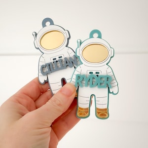 May include: Two astronaut-shaped name tags. The tags are white with blue and gold accents. One tag has the name "GITTAN" and the other has "RYDER". The astronaut helmets have gold visors. The tags are held in a hand against a white background.