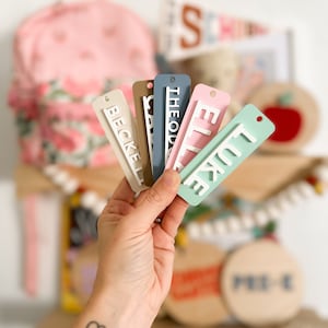 Op de afbeelding: Een hand houdt zes kleurrijke gepersonaliseerde naamstickers vast met witte letters. De namen zijn "Becke", "Cass", "Theodo", "Ellie", "Luke" en "Pre-K".