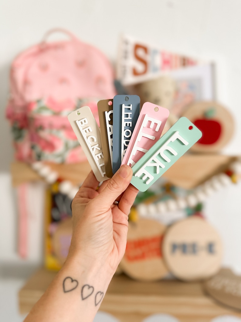 Op de afbeelding: Een hand houdt zes kleurrijke gepersonaliseerde naamstickers vast met witte letters. De namen zijn "Becke", "Cass", "Theodo", "Ellie", "Luke" en "Pre-K".