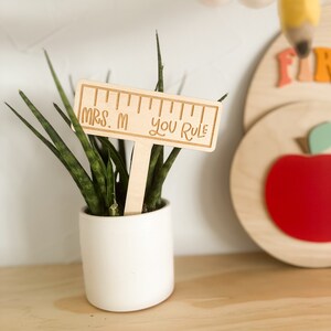 May include: Wooden ruler with the text "Mrs. M You Rule" on a wooden stick, inserted into a potted plant.