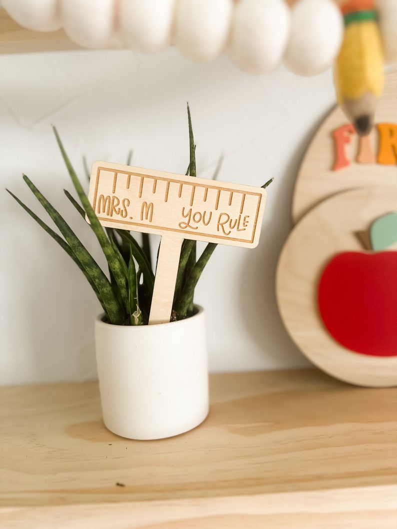 May include: Wooden ruler with the text "Mrs. M You Rule" on a wooden stick, inserted into a potted plant.
