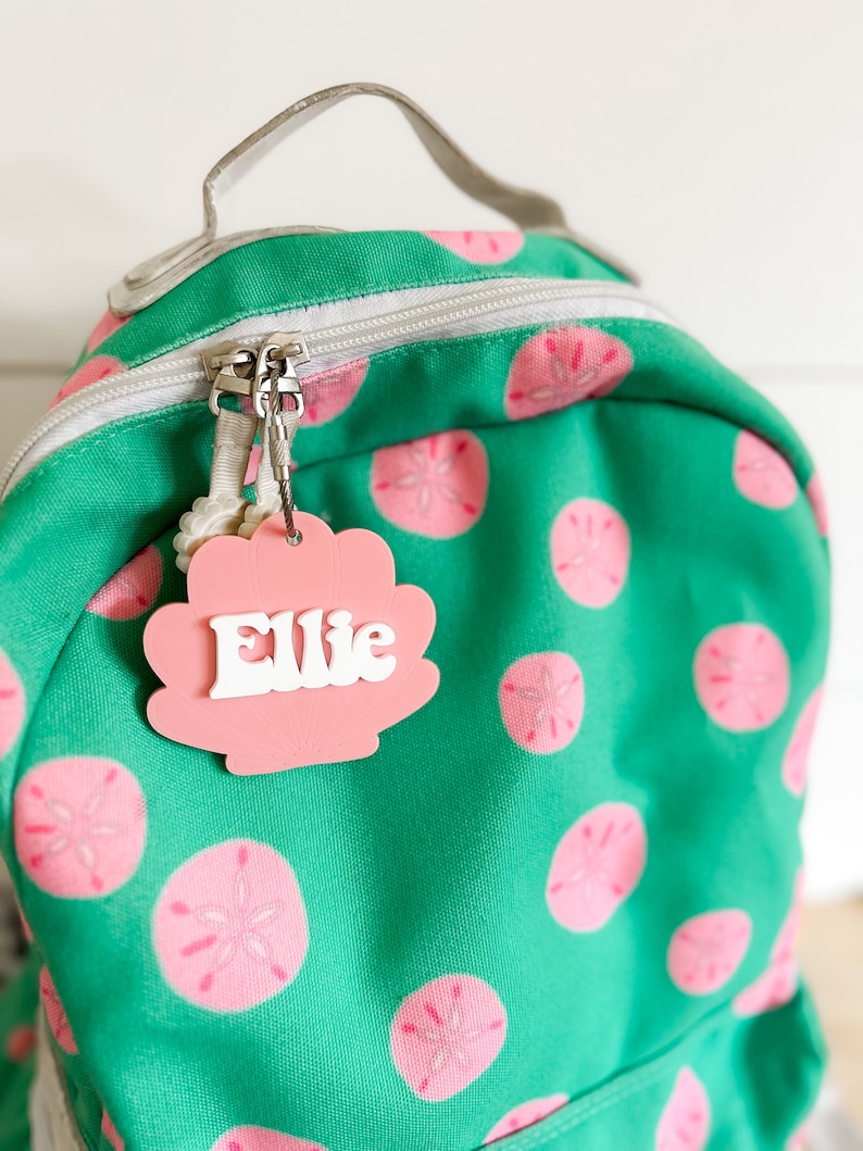 May include: A green backpack with a pink seashell pattern. A pink seashell name tag with the name "Ellie" is attached to the zipper.