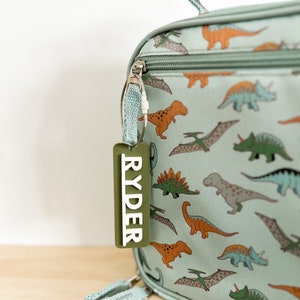 Op de afbeelding: Een lichtblauwe lunchtas met een dinosaurusprint en een groen gepersonaliseerd label met de tekst "RYDER".