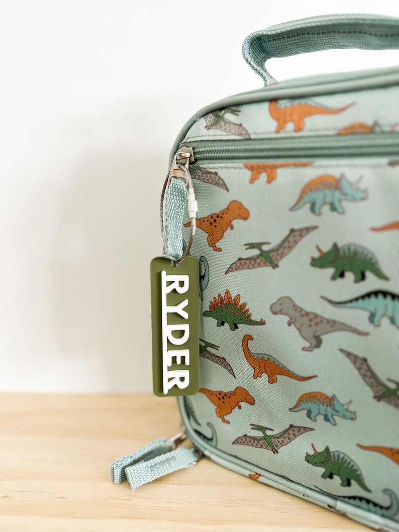 Op de afbeelding: Een lichtblauwe lunchtas met een dinosaurusprint en een groen gepersonaliseerd label met de tekst "RYDER".