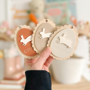 May include: Three wooden Easter egg-shaped ornaments with bunny silhouettes and scalloped edges. The ornaments are painted in shades of brown, beige, and peach. The names "Charlie", "Shea", and "Anthony" are written on the ornaments.