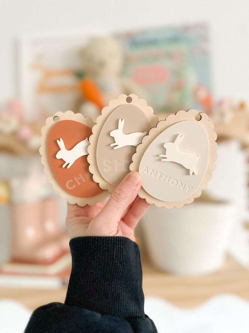 May include: Three wooden Easter egg-shaped ornaments with bunny silhouettes and scalloped edges. The ornaments are painted in shades of brown, beige, and peach. The names "Charlie", "Shea", and "Anthony" are written on the ornaments.