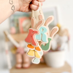 May include: Three wooden Easter ornaments shaped like letters, K, R, and J, with bunny ears. The letters are painted in pastel colors, including pink, blue, green, yellow, and orange.