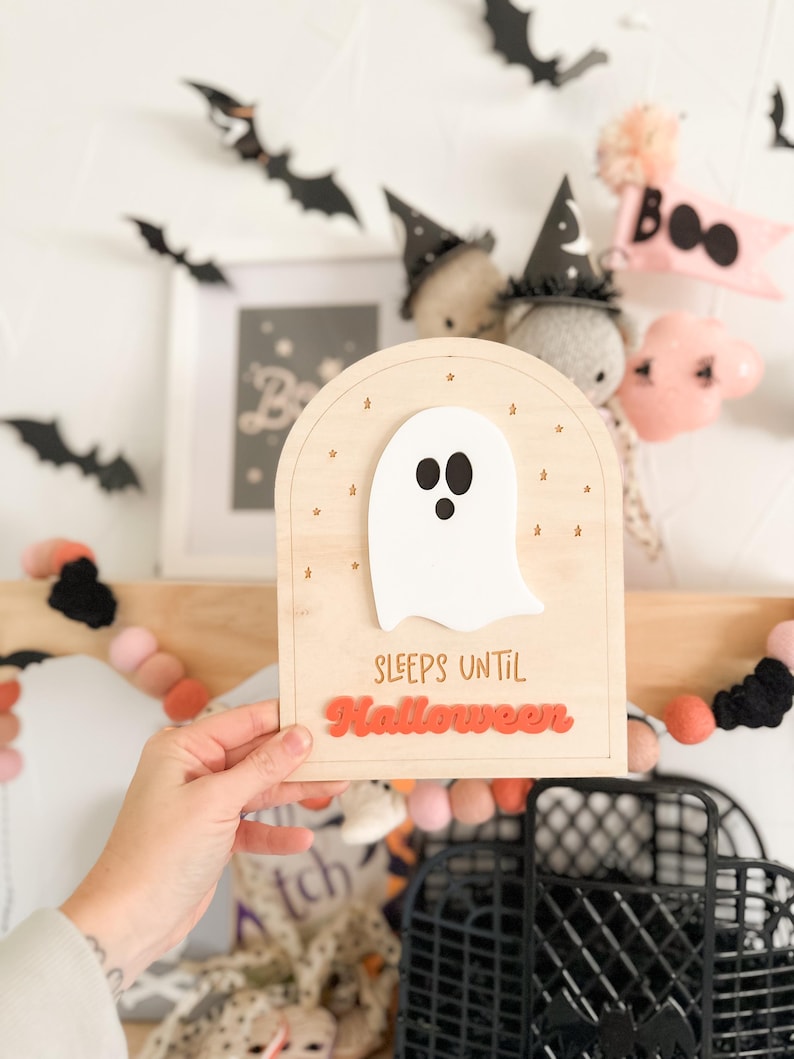 May include: A wooden sign with a white ghost and the text "Sleeps Until Halloween" in orange letters. The sign is decorated with small gold stars.