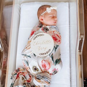 May include: A newborn baby girl swaddled in a floral swaddle blanket with a wooden name tag that reads "Charlotte Grey".