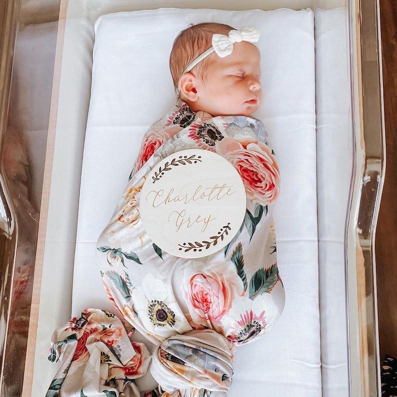 May include: A newborn baby girl swaddled in a floral swaddle blanket with a wooden name tag that reads "Charlotte Grey".