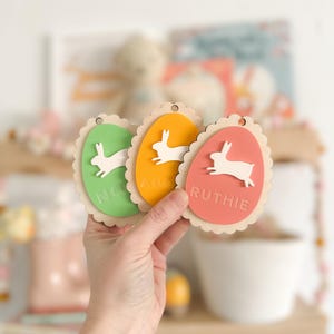 May include: Three wooden Easter egg ornaments with bunny cutouts and names "Nora", "Ava", and "Ruthie" in white lettering.