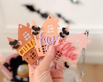 May include: Three house-shaped ornaments in shades of terracotta, peach, and light pink. Each ornament features cut-out windows, small black bat accents, and a personalized name in raised script lettering: Cillia, Faye, and Ellie.
