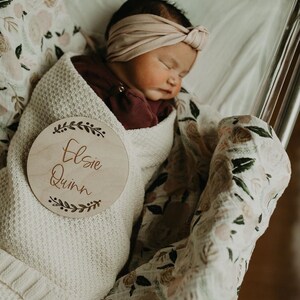 May include: A newborn baby girl is swaddled in a white blanket with a floral pattern. A wooden circle with the name "Elsie Quinn" is placed on the blanket. The baby is wearing a burgundy shirt and a light pink headband.