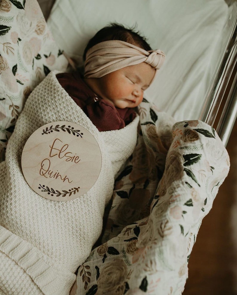 May include: A newborn baby girl is swaddled in a white blanket with a floral pattern. A wooden circle with the name "Elsie Quinn" is placed on the blanket. The baby is wearing a burgundy shirt and a light pink headband.