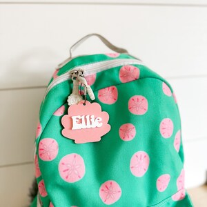 May include: A green backpack with a pink seashell-shaped name tag that says "Ellie" on it. The backpack has a white zipper and a white strap. The backpack is covered in a pattern of pink seashells.
