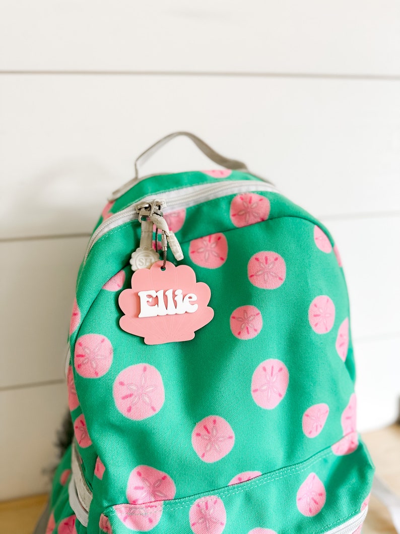 May include: A green backpack with a pink seashell-shaped name tag that says "Ellie" on it. The backpack has a white zipper and a white strap. The backpack is covered in a pattern of pink seashells.