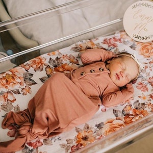 May include: A newborn baby sleeping in a clear plastic bassinet with a floral patterned sheet. The baby is wearing a peach-colored knotted gown and a headband. A wooden sign with the name "Emma Mae" is hanging above the bassinet.