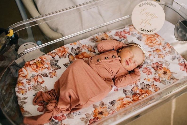 May include: A newborn baby sleeping in a clear plastic bassinet with a floral patterned sheet. The baby is wearing a peach-colored knotted gown and a headband. A wooden sign with the name "Emma Mae" is hanging above the bassinet.