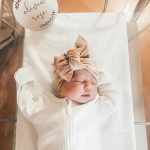 May include: A newborn baby wearing a white onesie and a brown velvet bow headband. A wooden sign with the text "Olivia Rose" is in the background.