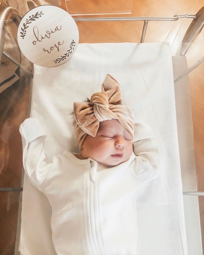 May include: A newborn baby wearing a white onesie and a brown velvet bow headband. A wooden sign with the text "Olivia Rose" is in the background.