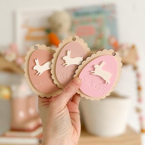 May include: Three pink and white wooden Easter egg ornaments with bunny silhouettes and the names "Colin", "Gi", and "Sage" written on them.