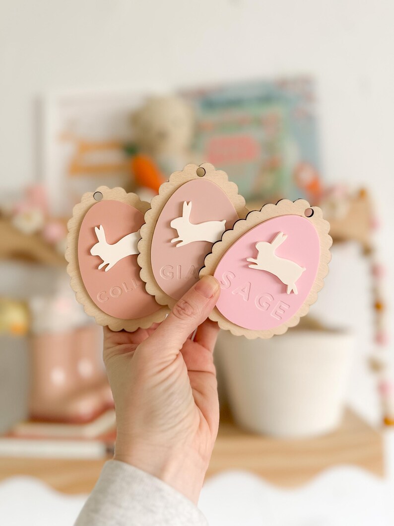 May include: Three pink and white wooden Easter egg ornaments with bunny silhouettes and the names "Colin", "Gi", and "Sage" written on them.