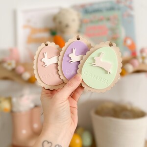 May include: Three pastel-colored wooden Easter egg ornaments with scalloped edges. Each egg features a white bunny silhouette and a name: "Casey", "Cam", and "Gambel".