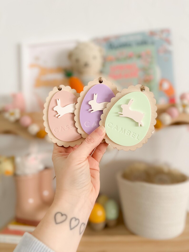 May include: Three pastel-colored wooden Easter egg ornaments with scalloped edges. Each egg features a white bunny silhouette and a name: "Casey", "Cam", and "Gambel".