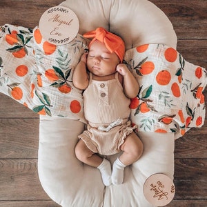 May include: A baby girl wearing a peach-colored headband and a beige ribbed outfit is lying in a white oval-shaped baby nest. The baby is surrounded by a white and orange patterned muslin swaddle. Two wooden signs with the text "Abigail Eve" and "one month" are visible in the image.