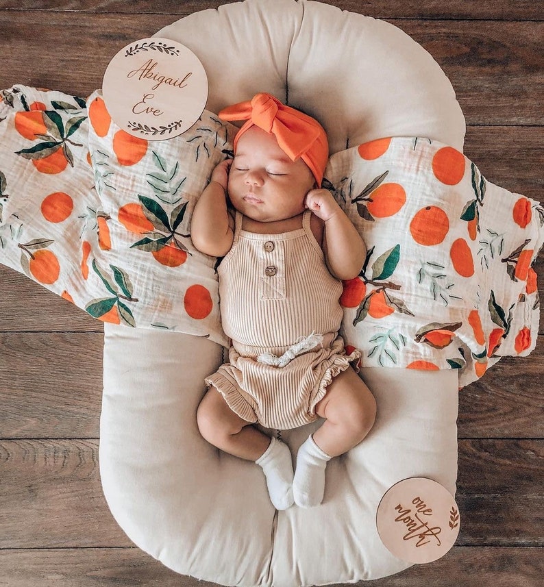 May include: A baby girl wearing a peach-colored headband and a beige ribbed outfit is lying in a white oval-shaped baby nest. The baby is surrounded by a white and orange patterned muslin swaddle. Two wooden signs with the text "Abigail Eve" and "one month" are visible in the image.