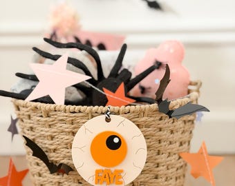 May include: A woven tan basket decorated with a round white and orange eyeball ornament featuring the name FAYE in orange letters. The basket is adorned with black bat cutouts and orange and pink star shapes, with more bats visible above.