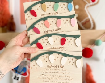 May include: Wooden Christmas cards with colorful light bulb designs. Each card is personalized with names and a holiday message. The cards are held in a hand, with a gingerbread man and decorative trees in the background.
