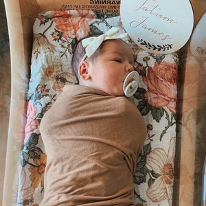 May include: A newborn baby girl wearing a white headband and a brown swaddle blanket is lying on a white and floral patterned sheet. A wooden sign with the name "Tatum James" is visible in the background.