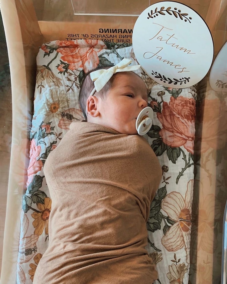 May include: A newborn baby girl wearing a white headband and a brown swaddle blanket is lying on a white and floral patterned sheet. A wooden sign with the name "Tatum James" is visible in the background.