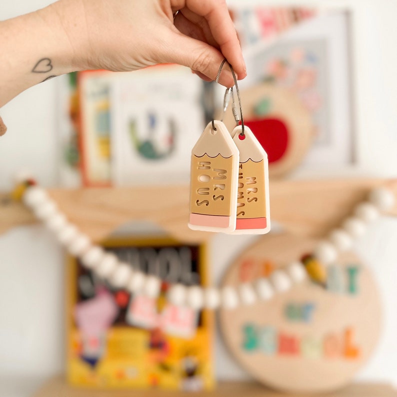 Puede incluir: Dos llaveros en forma de l&aacute;piz amarillo con gomas de borrar rosas. Los llaveros est&aacute;n personalizados con el texto "Mrs. Thomas" y "Mrs. Hous".