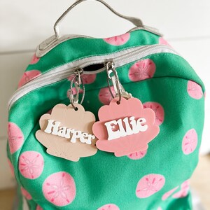May include: Two pink and beige shell-shaped name tags with the names "Harper" and "Ellie" written in white letters. The tags are attached to a green backpack with a white zipper and pink and white sand dollar designs.