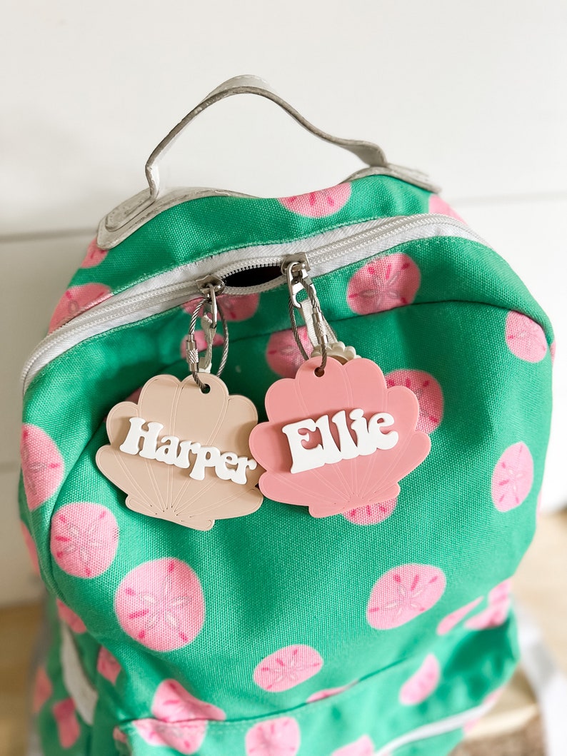 May include: Two pink and beige shell-shaped name tags with the names "Harper" and "Ellie" written in white letters. The tags are attached to a green backpack with a white zipper and pink and white sand dollar designs.