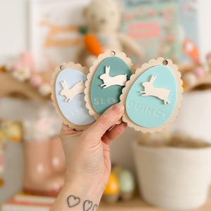 May include: Three wooden Easter egg-shaped ornaments with bunny silhouettes and scalloped edges. The ornaments are painted in light blue, teal, and light green. The names "Harper", "Sloan", and "Quincy" are written on the ornaments.