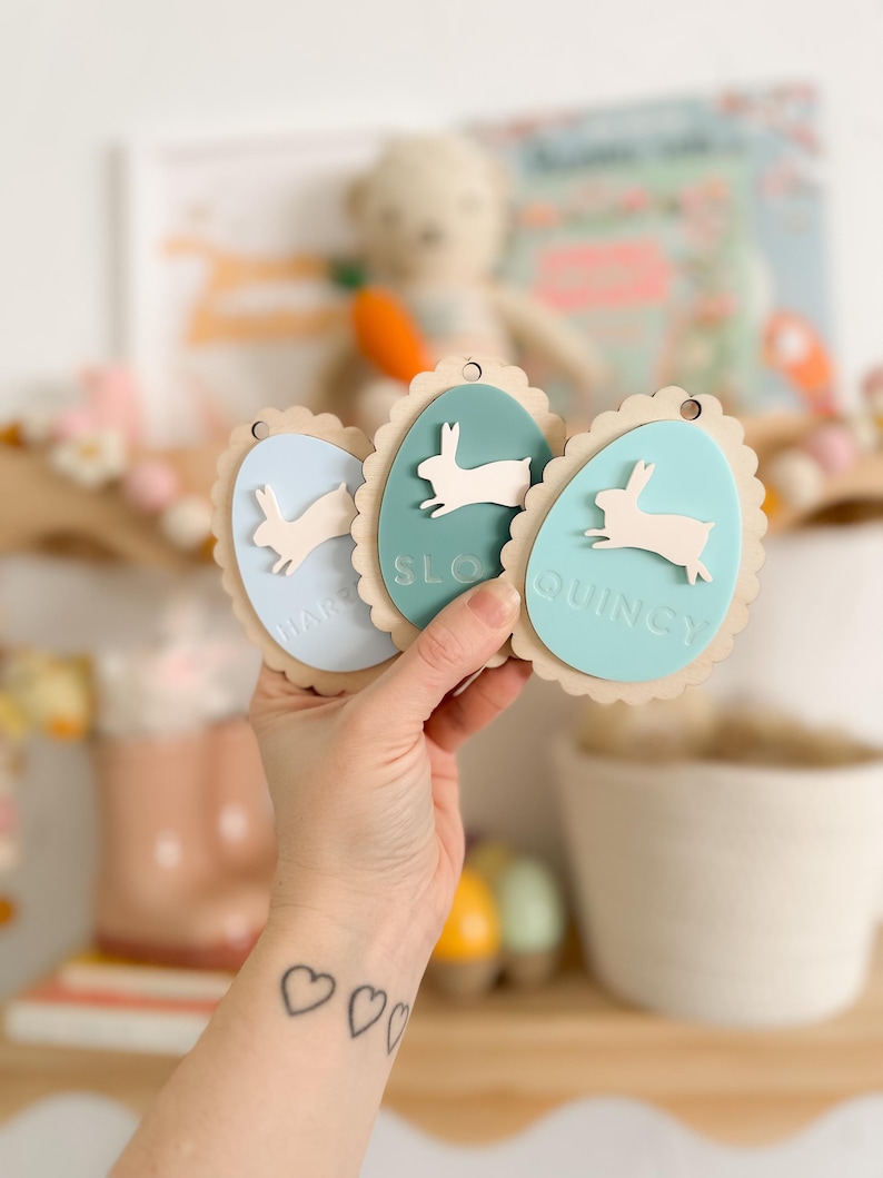 May include: Three wooden Easter egg-shaped ornaments with bunny silhouettes and scalloped edges. The ornaments are painted in light blue, teal, and light green. The names "Harper", "Sloan", and "Quincy" are written on the ornaments.