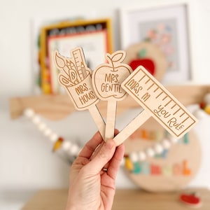 May include: Three wooden teacher appreciation signs with a pencil and paintbrush design, an apple design, and a ruler design. The signs read "Mrs. Hous", "Mrs. Gentil", and "Mrs. M You Rule".