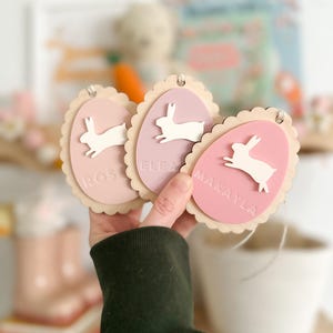 May include: Three pink and white wooden Easter egg ornaments with bunny silhouettes and the names "Rosalee", "Eleanor", and "Makayla" written on them.