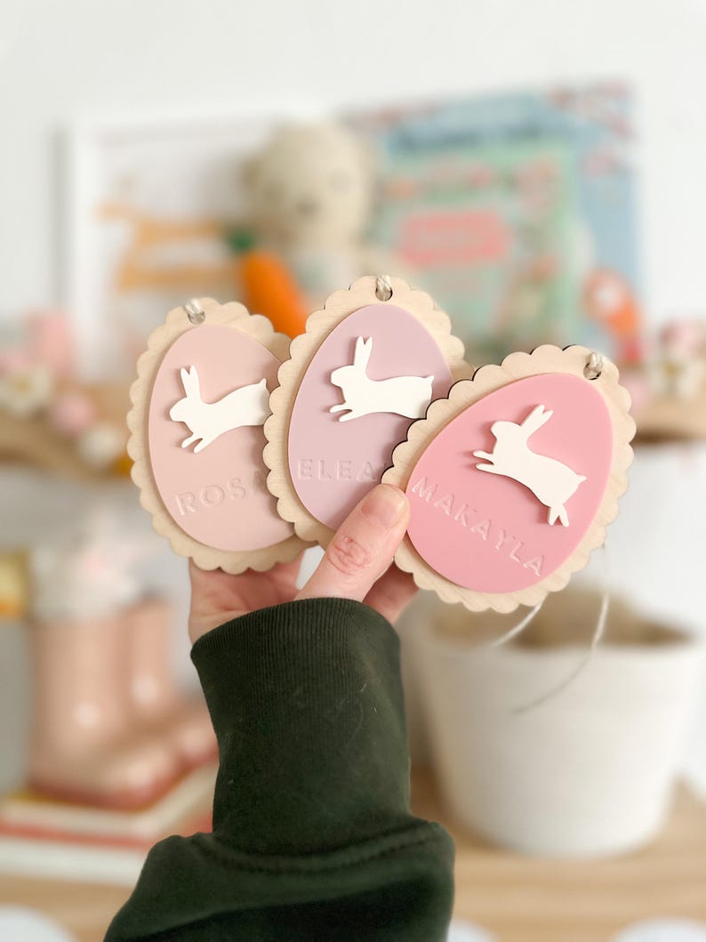 May include: Three pink and white wooden Easter egg ornaments with bunny silhouettes and the names "Rosalee", "Eleanor", and "Makayla" written on them.