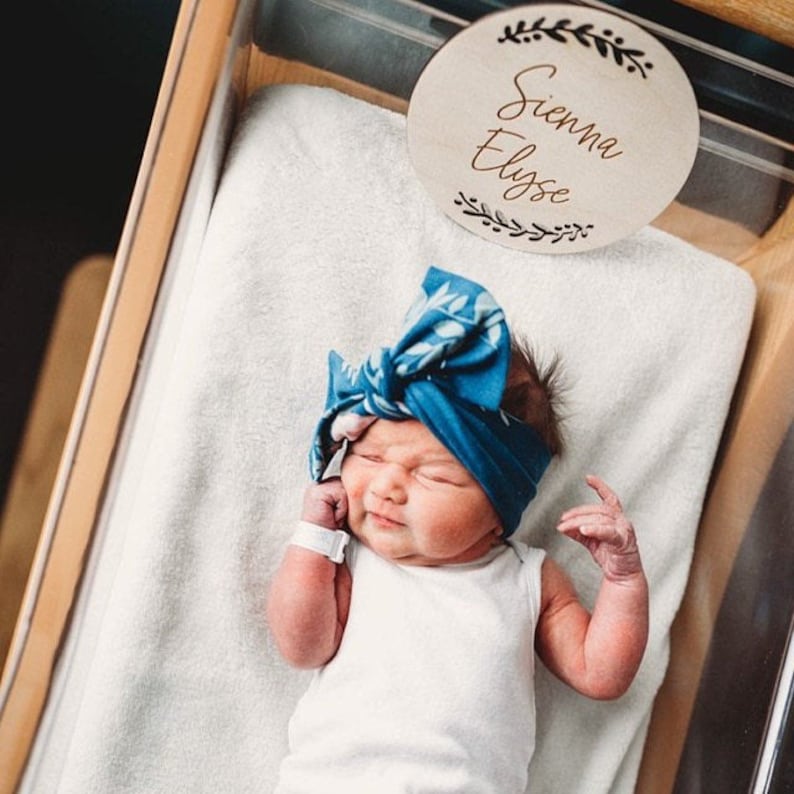 May include: A newborn baby girl wearing a blue and white headband with a floral pattern, lying in a white bassinet. A wooden sign with the name "Sienna Elyse" is above the baby.