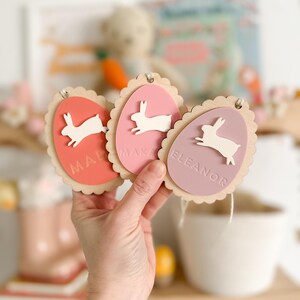 May include: Three wooden Easter egg ornaments with bunny cutouts and personalized names. The eggs are painted in shades of pink and red.