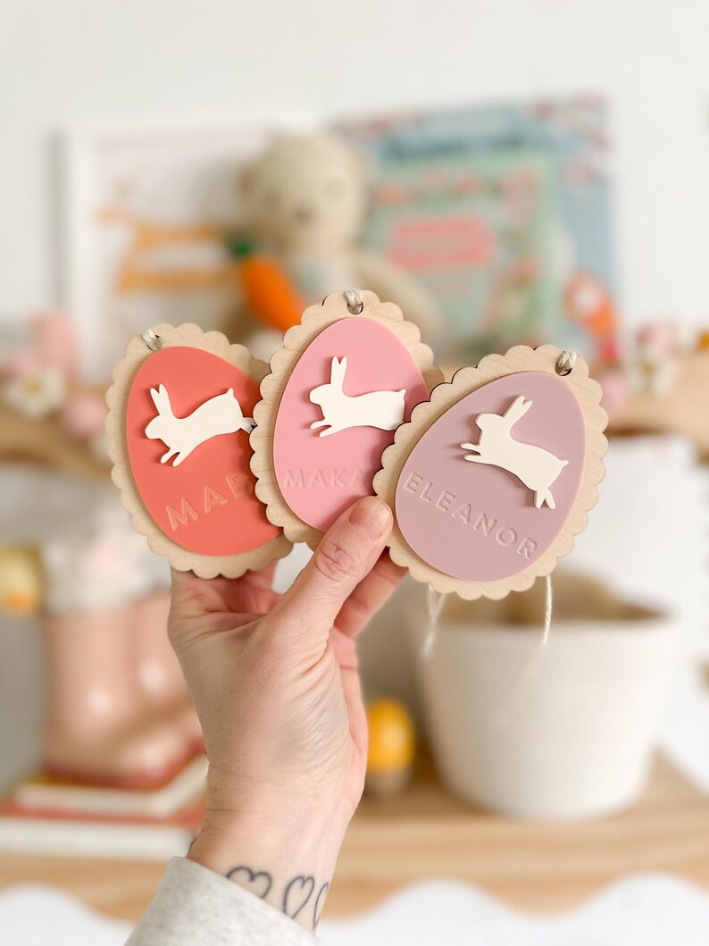 May include: Three wooden Easter egg ornaments with bunny cutouts and personalized names. The eggs are painted in shades of pink and red.