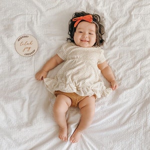 May include: A baby girl wearing a white dress with brown polka dots and a red bow headband. She is lying on a white blanket and holding a wooden sign that says "Lilah Joy".