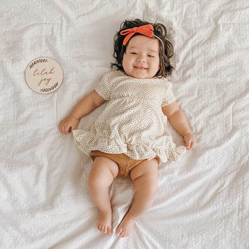 May include: A baby girl wearing a white dress with brown polka dots and a red bow headband. She is lying on a white blanket and holding a wooden sign that says "Lilah Joy".